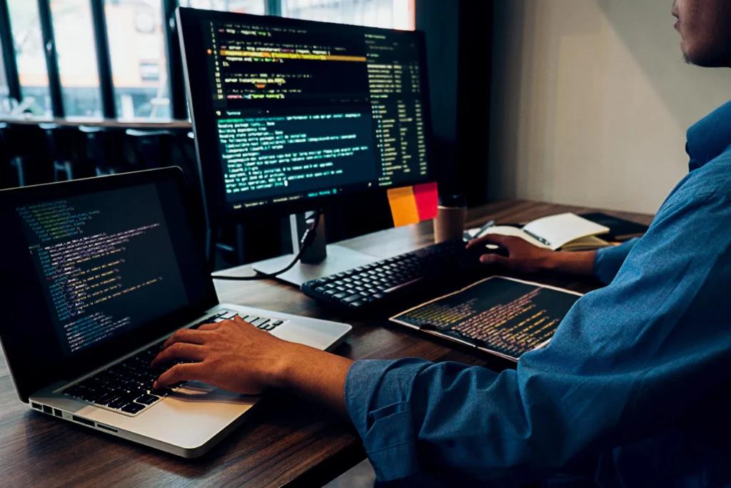 Person working on multiple screens displaying code in a dimly lit office, creating a focused and productive atmosphere.