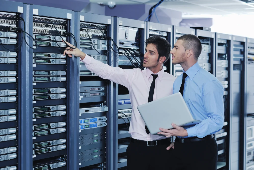Two IT professionals in shirts and ties examine server racks in a data center. One points at cables, while the other holds a laptop, appearing focused.